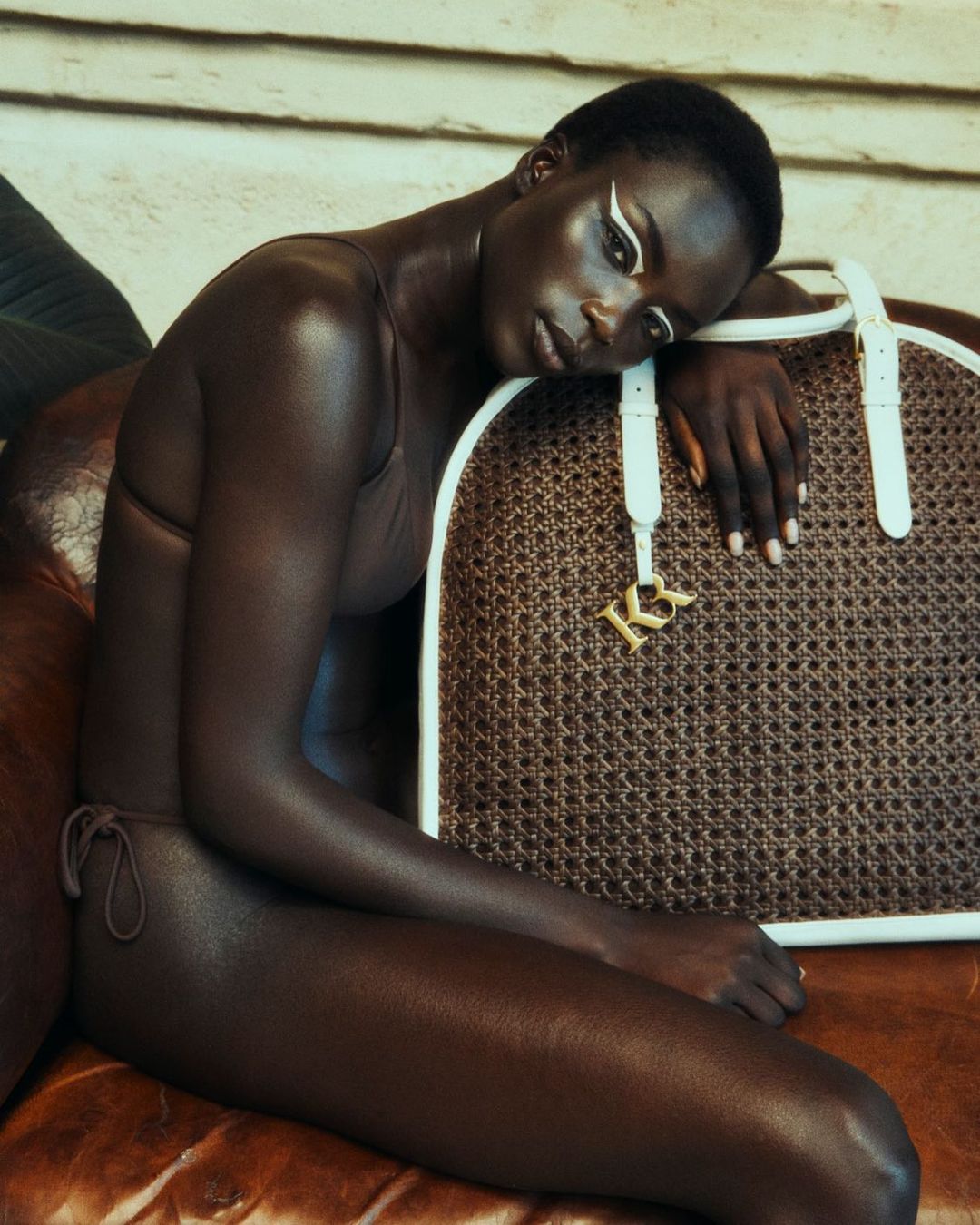 Elegant dark-skinned woman poses with a brown woven handbag, showcasing white eyeliner and minimalist fashion in a soft, inviting light.