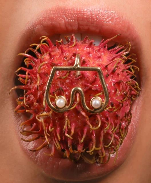 Close-up shot of a person's mouth holding a rambutan fruit with a gold pendant featuring pearls, highlighting a beauty and fashion concept.