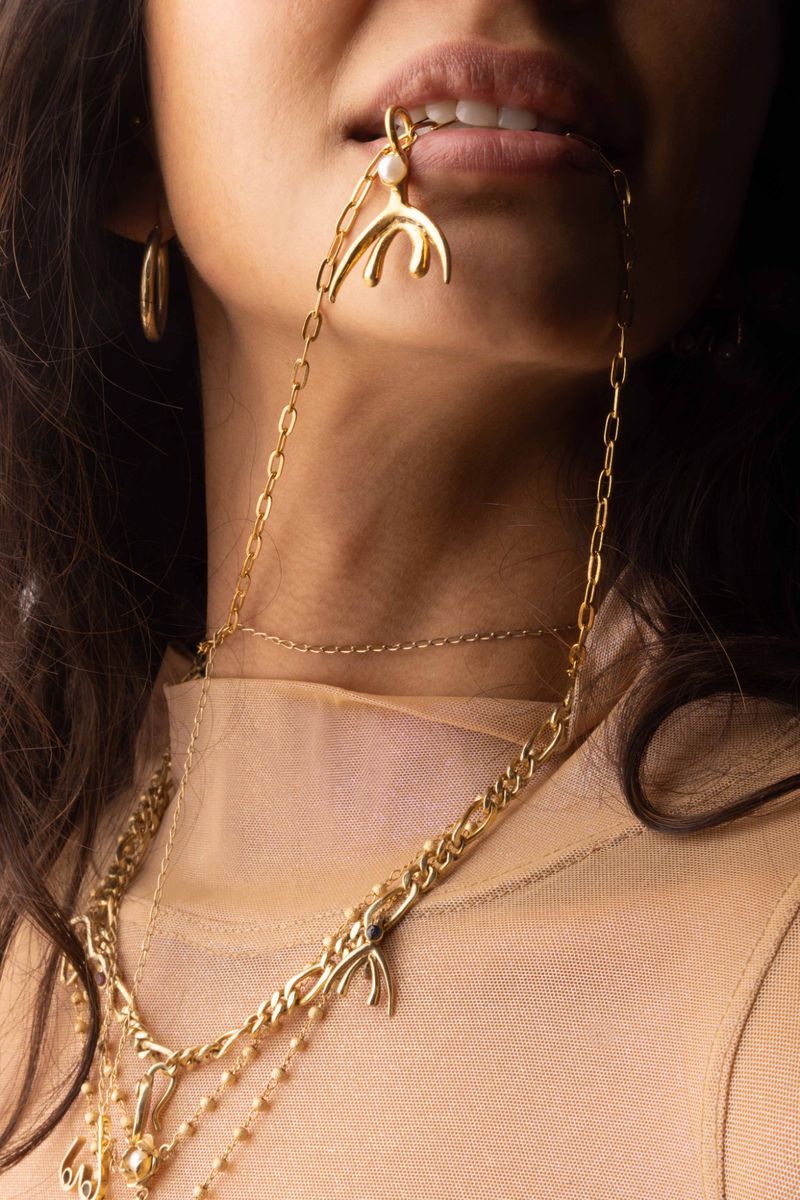 Close-up shot of a woman wearing gold jewelry, including a chunky necklace, a chain necklace, and earrings, against a dark background.