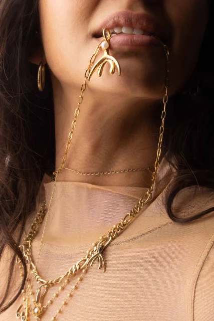 Close-up shot of a woman wearing gold jewelry, including a chunky necklace, a chain necklace, and earrings, against a dark background.