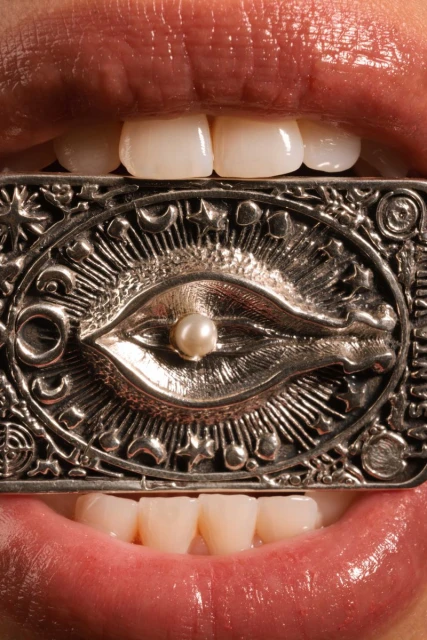 Close-up of an adult woman's mouth with white teeth. She holds a metal talisman in her mouth. The talisman has an eye design with a pearl.