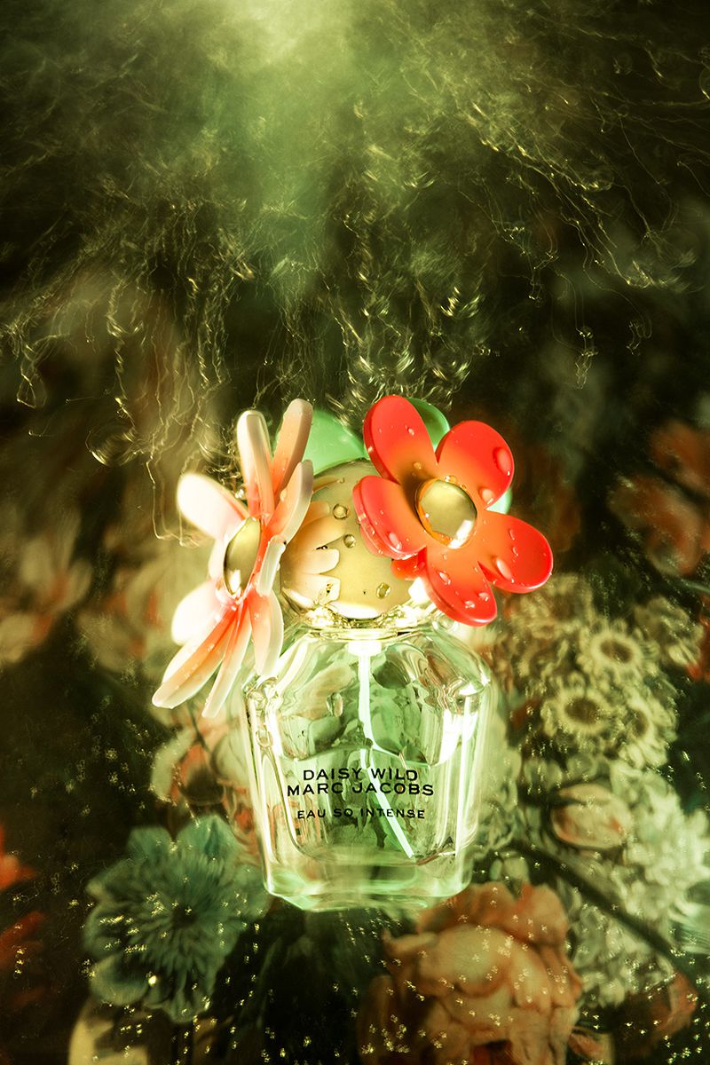 Close-up product shot of clear glass Marc Jacobs perfume bottle with flower-shaped cap, set against a background of floral patterns.