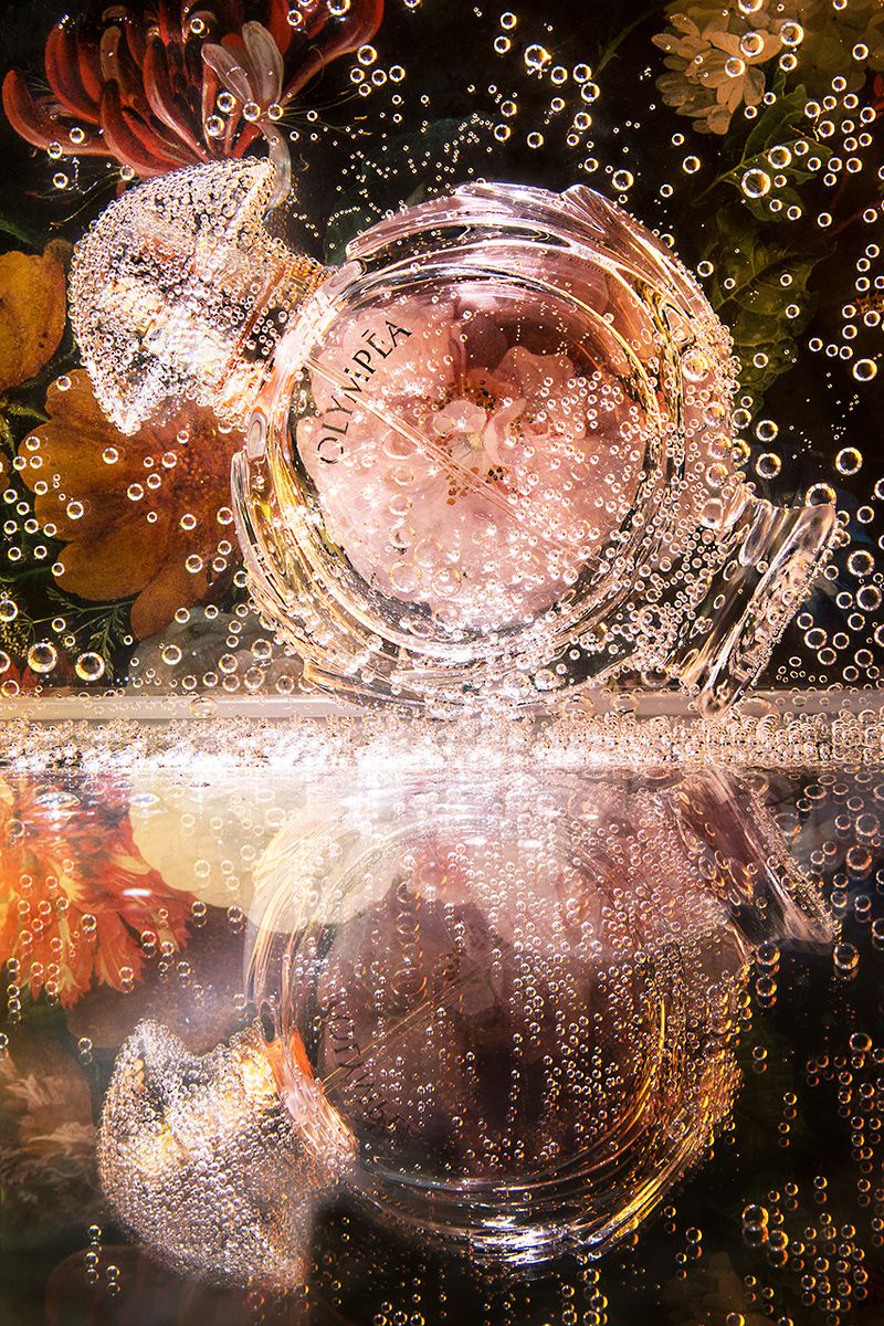 Perfume bottle submerged in water surrounded by flowers, creating a surreal and artistic composition with dark background.