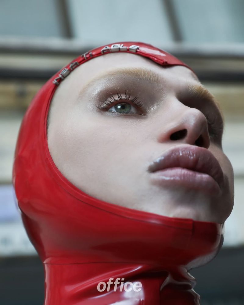 Close-up of a woman's face wearing a red latex hood, shot from a low angle indoors. The hood has silver text across the forehead.