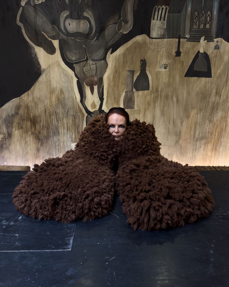 A woman poses indoors, partially obscured by brown, wooly objects, against a backdrop with painted figures and architectural elements.