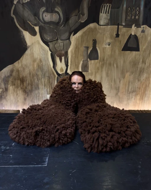 A woman poses indoors, partially obscured by brown, wooly objects, against a backdrop with painted figures and architectural elements.