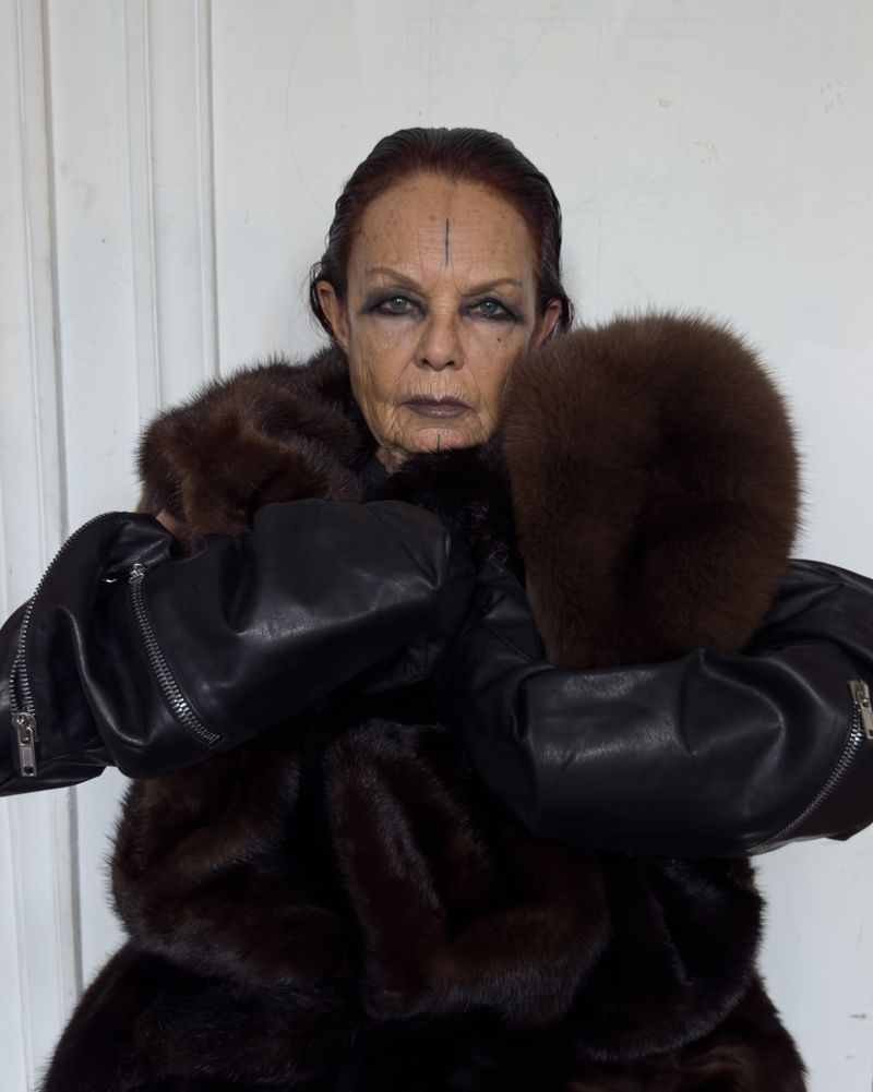 Adult female with shaved head wears dark eye makeup, an unusual head marking, fur coat, and leather jacket against a neutral wall.