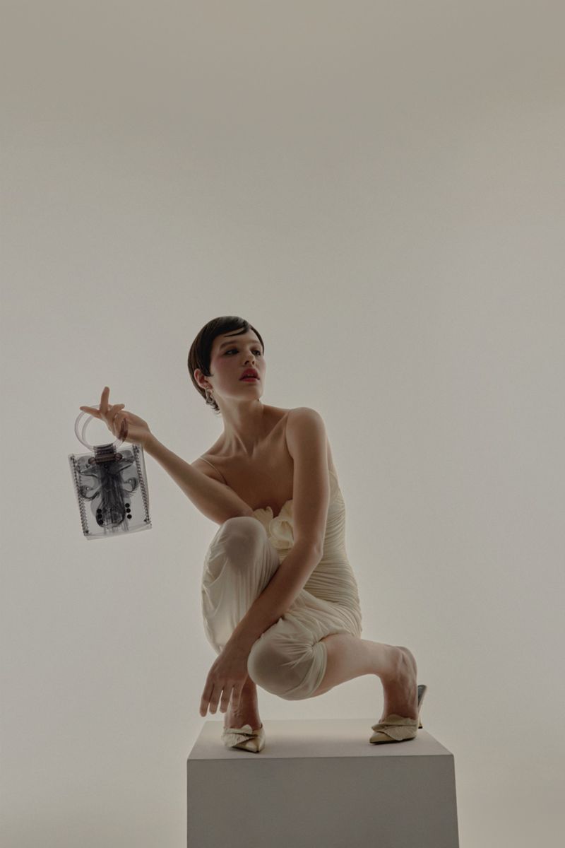 Fashionable woman with short hair in stylish white dress poses with a clear, sculptural handbag while kneeling in a bright studio.
