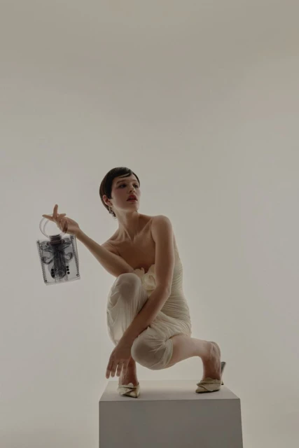 Fashionable woman with short hair in stylish white dress poses with a clear, sculptural handbag while kneeling in a bright studio.