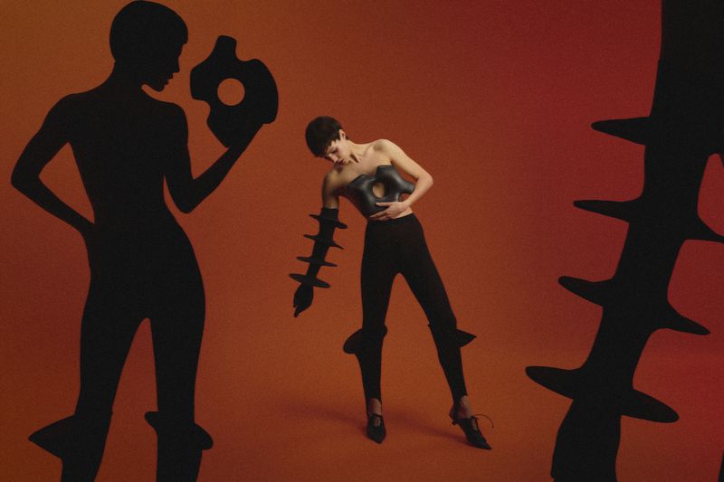 Artistic fashion photo featuring a female model in black, holding a geometric object against a gradient orange background with silhouettes.