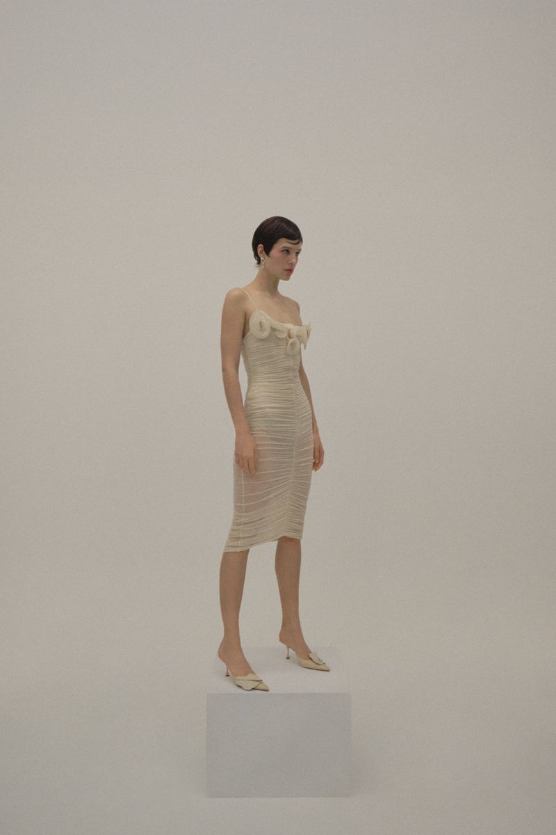 A slender woman with short dark hair poses in a cream-colored midi dress and heels on a white cube against a seamless backdrop.
