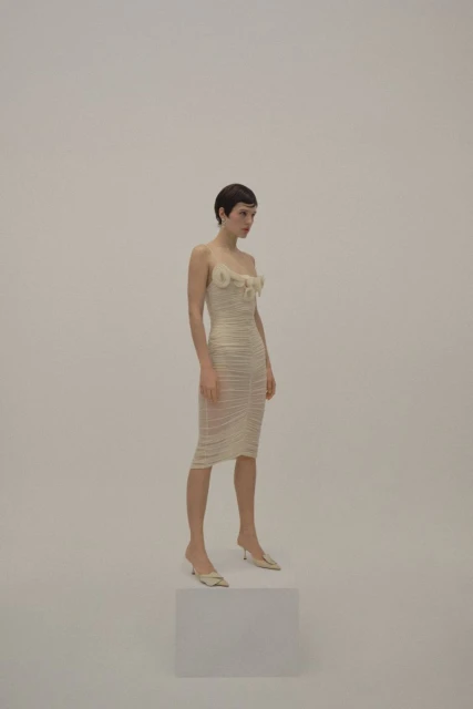 A slender woman with short dark hair poses in a cream-colored midi dress and heels on a white cube against a seamless backdrop.