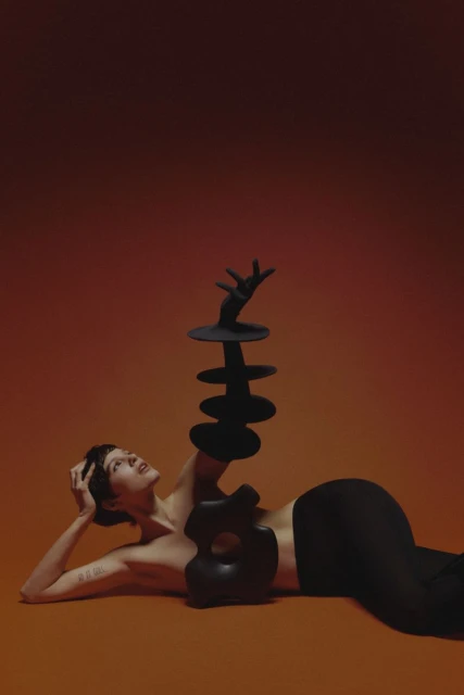 Woman poses with artistic sculpture on the floor, wearing fitted black clothing against warm orange and reddish-brown backdrop in photo studio.