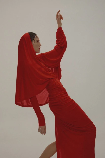 A woman in a red dress and head covering poses with her arm raised against a light background in a fashion editorial.
