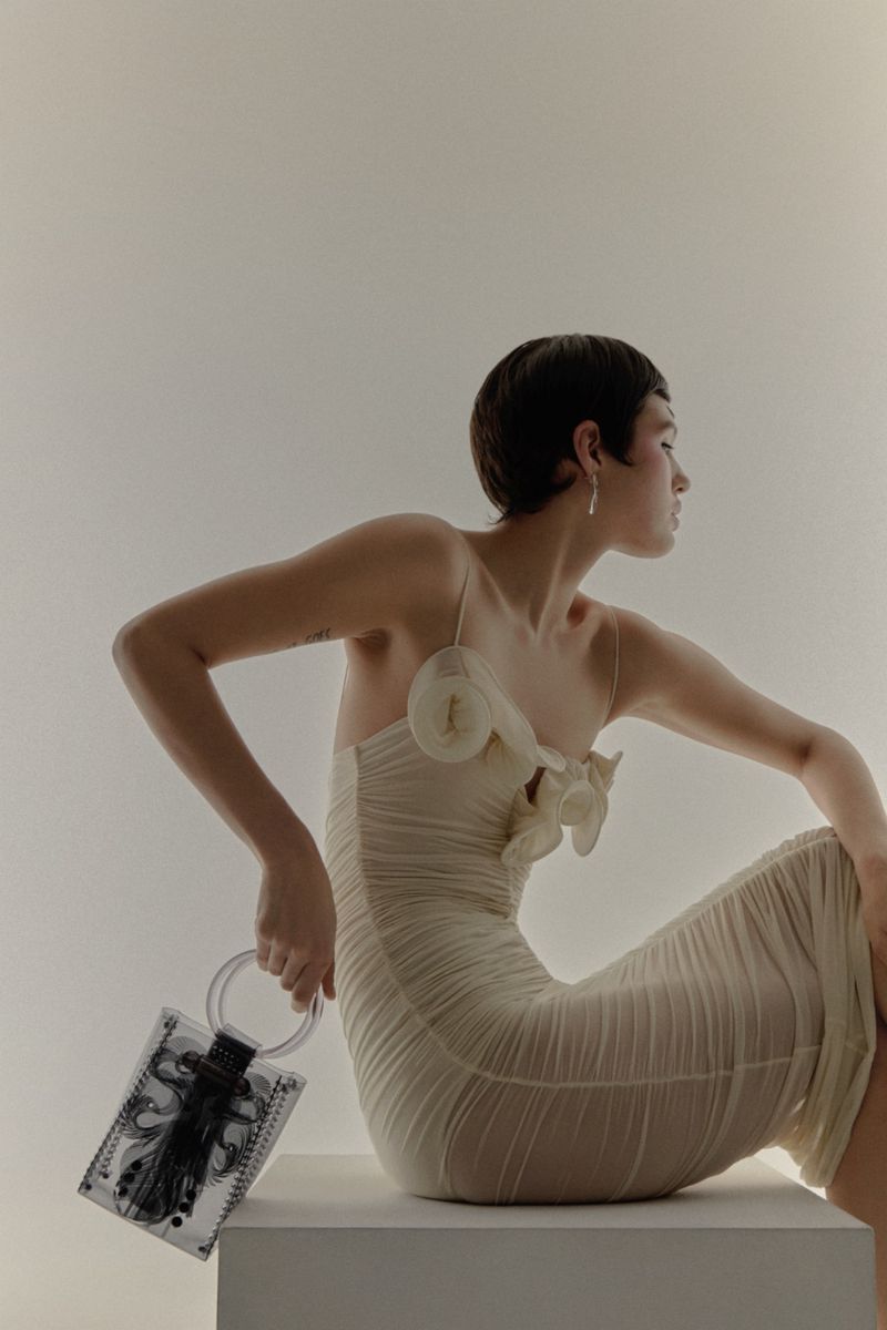 Woman in a light yellow dress sitting indoors with a transparent black-and-white purse in hand, captured in a studio setting.