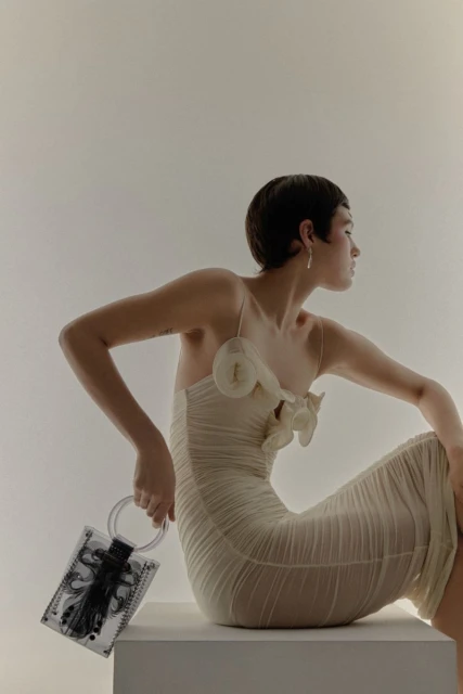 Woman in a light yellow dress sitting indoors with a transparent black-and-white purse in hand, captured in a studio setting.