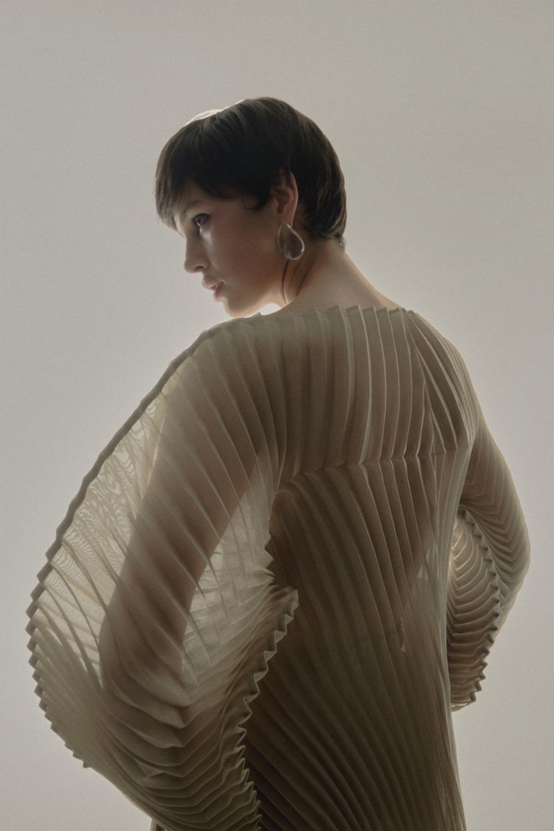 Profile shot of an adult woman with short brown hair wearing a pleated top and statement earrings in front of a neutral backdrop.