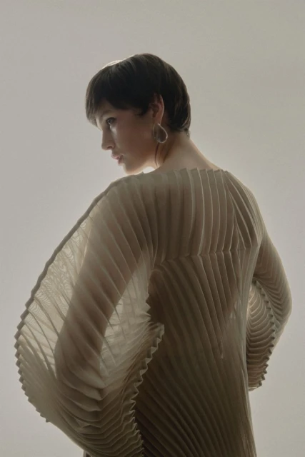 Profile shot of an adult woman with short brown hair wearing a pleated top and statement earrings in front of a neutral backdrop.