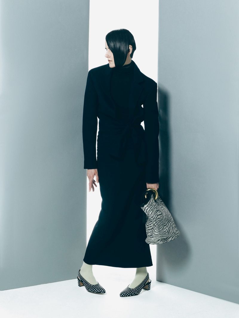 A slender woman in a black dress and polka dot heels poses between gray walls, holding a zebra print bag, looking off to the left.