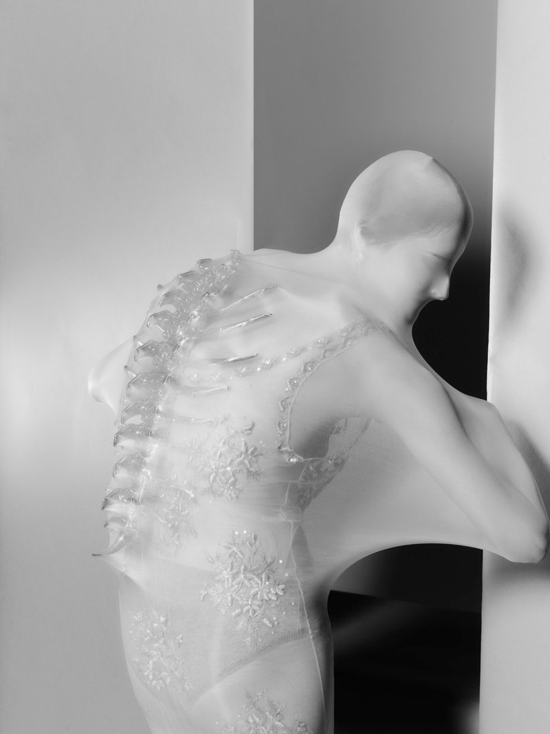 Ghostly pale mannequin draped in transparent white fabric with snowflake details, posed indoors in a corner. 