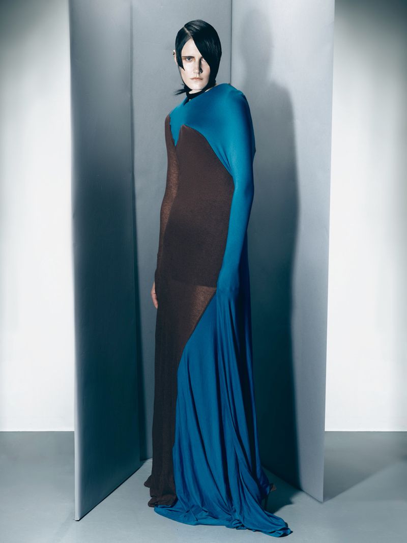 Fashion model poses in a full-length draped brown and blue gown against a graduated grey backdrop in a studio setting with diffused lighting.