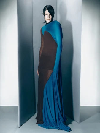 Fashion model poses in a full-length draped brown and blue gown against a graduated grey backdrop in a studio setting with diffused lighting.