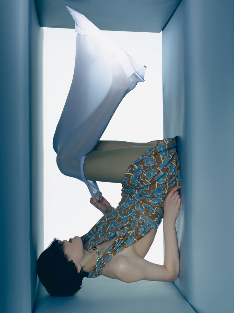 Artistic fashion shot of woman in patterned dress and stockings, positioned in a blue cube, surreal and visually striking studio portrait.