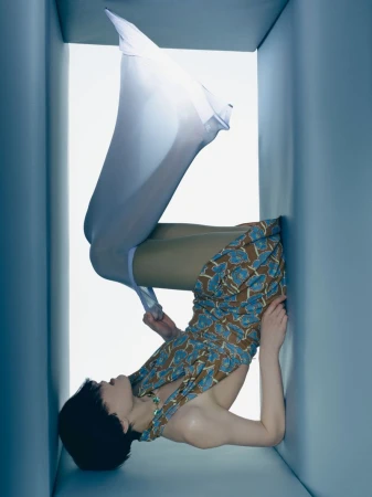 Artistic fashion shot of woman in patterned dress and stockings, positioned in a blue cube, surreal and visually striking studio portrait.