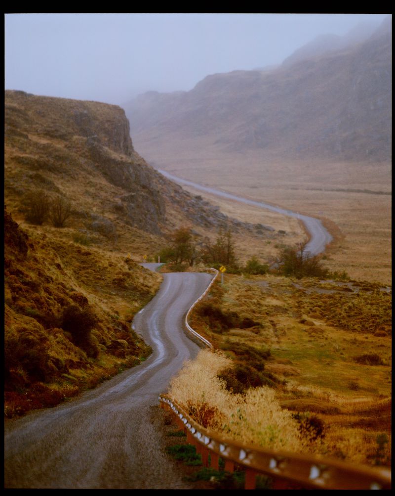 Winding asphalt road through a valley with golden fields, hills, and fog. Overcast daylight scene with guard rail on the roadside.