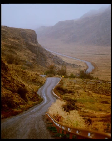 Winding asphalt road through a valley with golden fields, hills, and fog. Overcast daylight scene with guard rail on the roadside.