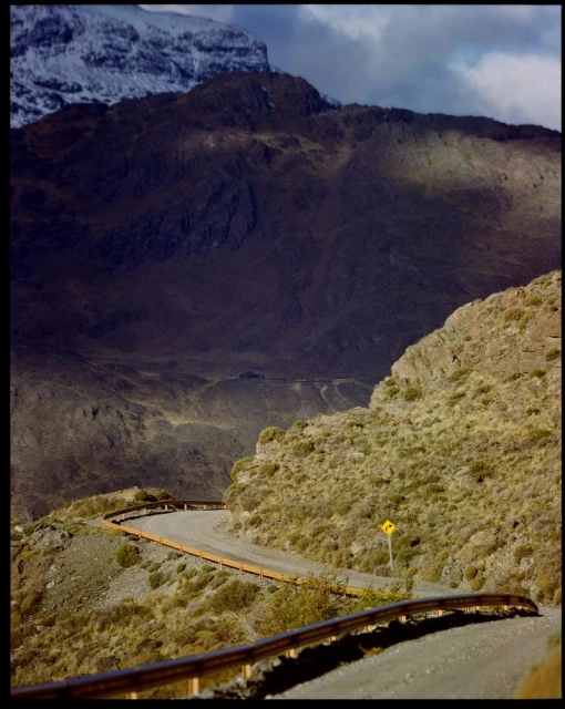 Winding road leads through a rugged mountain landscape on a sunny day, with snowy peaks in the distance, suggesting a remote and scenic location.