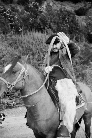 A man in a beret rides a horse, holding the reins and a rope, in a black and white outdoor setting with natural backgrounds.
