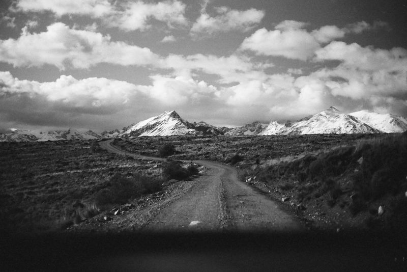 Monochrome landscape featuring a dirt road winding through grassy terrain toward snow-capped mountains under a cloudy sky.