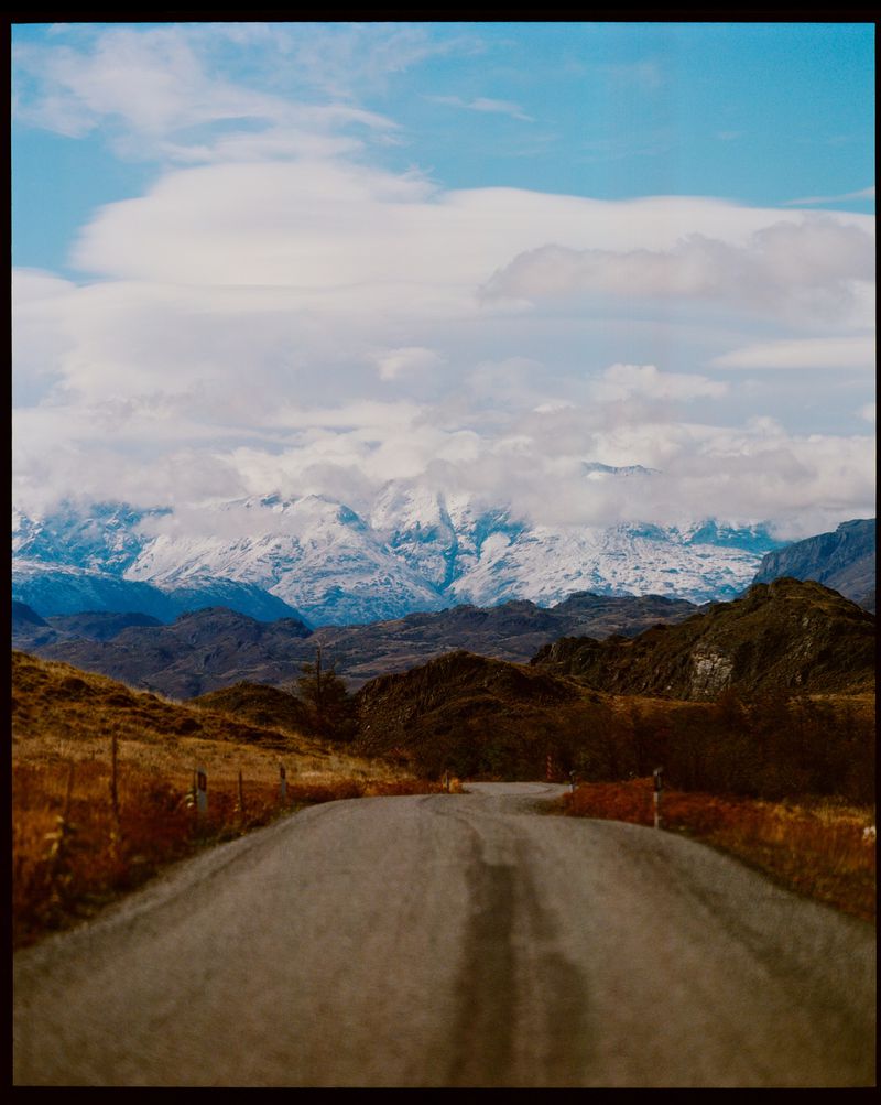 Scenic road leads to snow-covered mountains under a partly cloudy sky, highlighting natural beauty and a sense of adventure in a remote area.