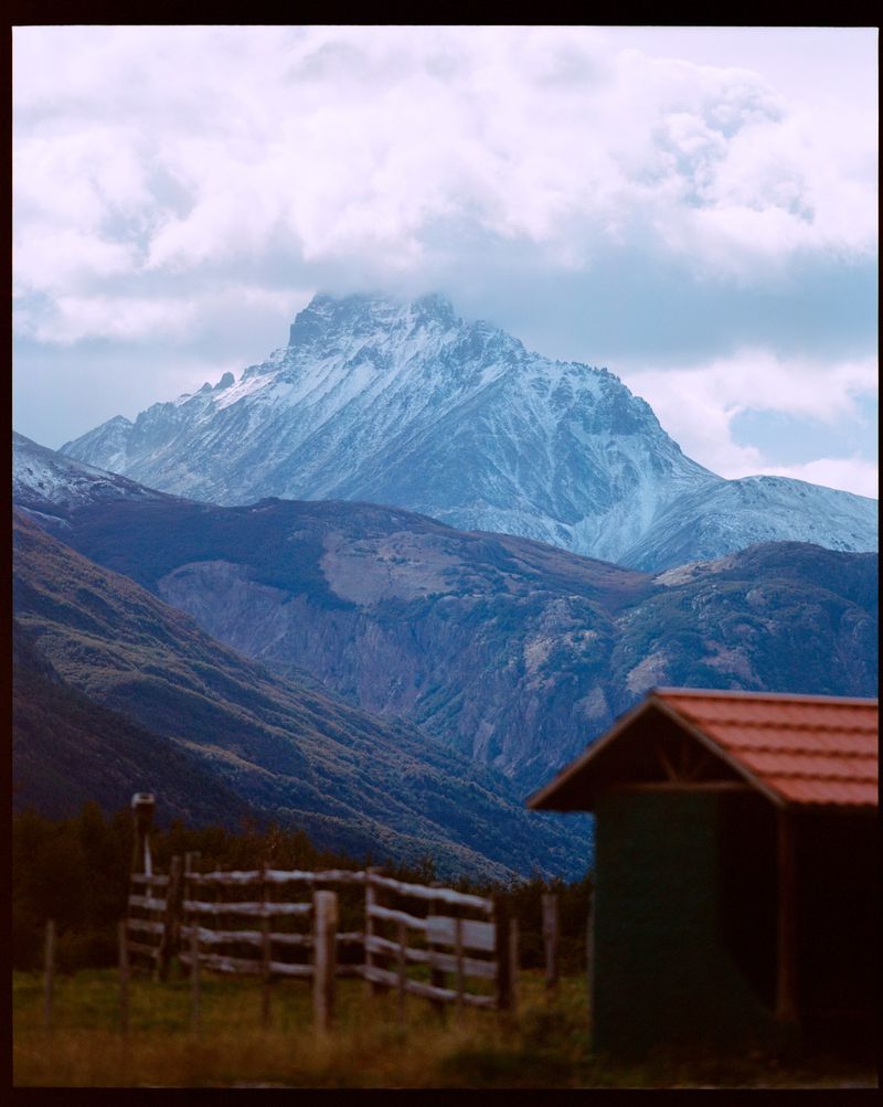 Scenic landscape featuring a snow-capped mountain range beneath cloudy skies, complemented by a wooden fence and a small shed in the foreground.