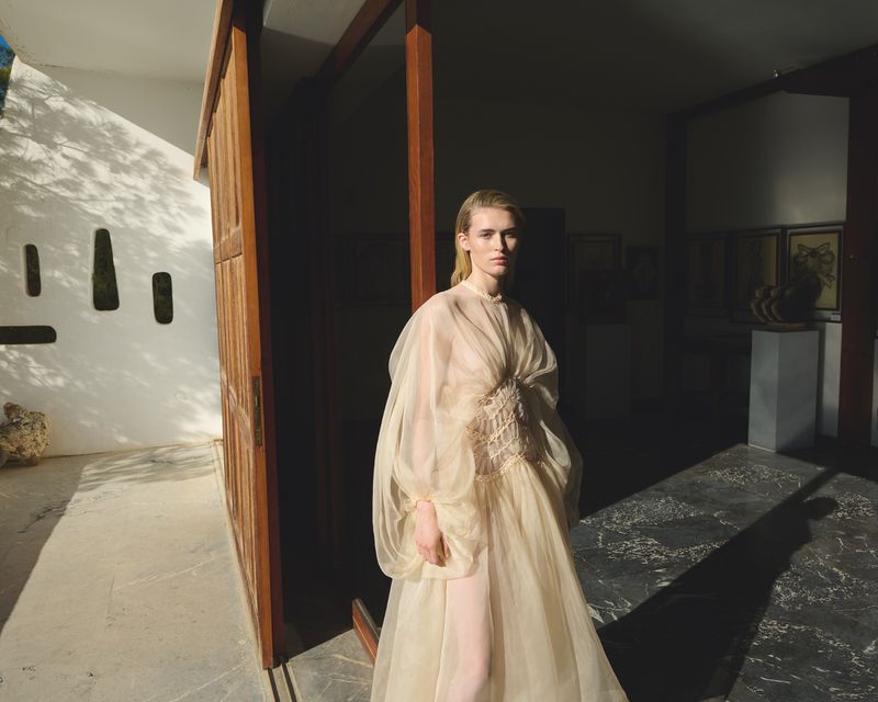 A slender woman in a sheer dress stands in a sunlit architectural space with modern design, creating a fashion-editorial-style image.