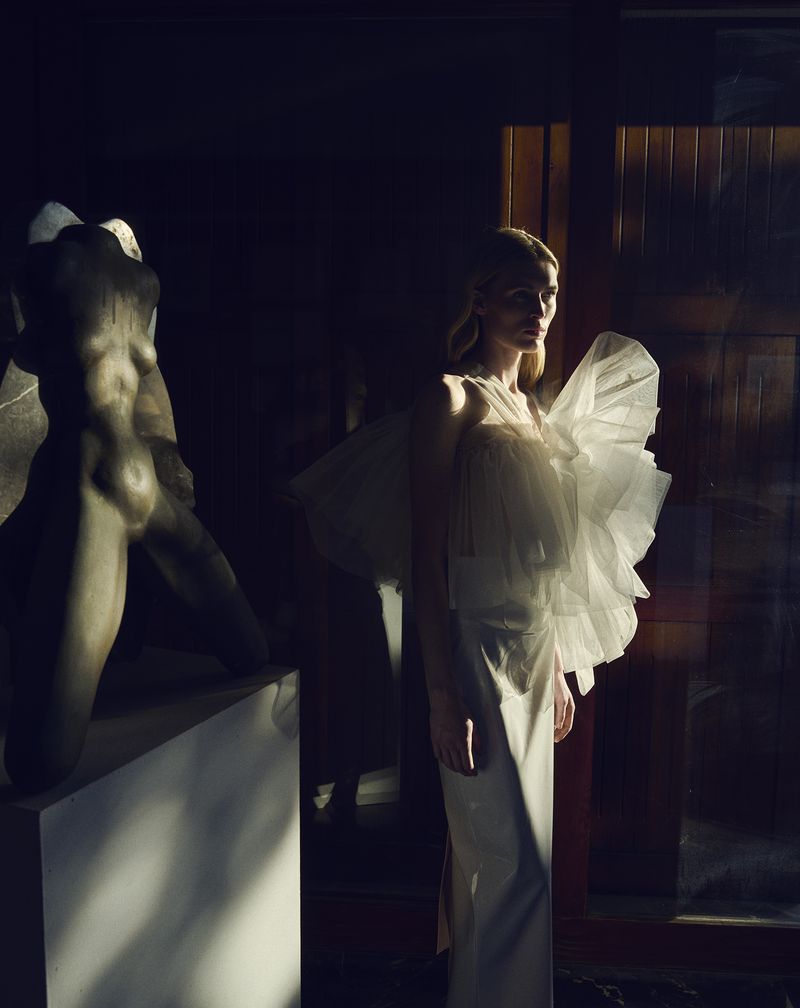 Woman in an off-white ruffled dress poses next to a classical sculpture in dramatic lighting. Fashion editorial shot.