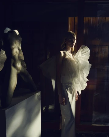 Woman in an off-white ruffled dress poses next to a classical sculpture in dramatic lighting. Fashion editorial shot.