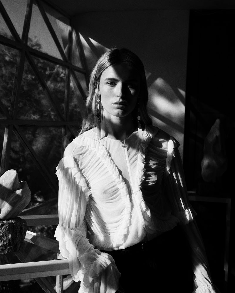 Black and white portrait of a woman with long hair wearing a ruffled blouse, standing indoors with filtered sunlight.