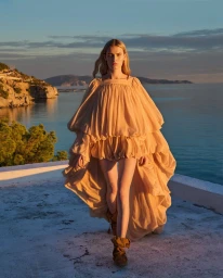 Model wearing a voluminous ruffled peach dress walking on a terrace overlooking the sea at sunset
