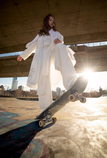 Woman in white outfit balances on skateboard under bridge with cityscape background and bright sunlight in fashion lifestyle shot.