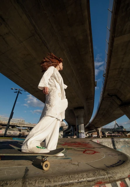 Young woman in white outfit skateboarding under a bridge against a blue sky. She has auburn hair and is looking away.