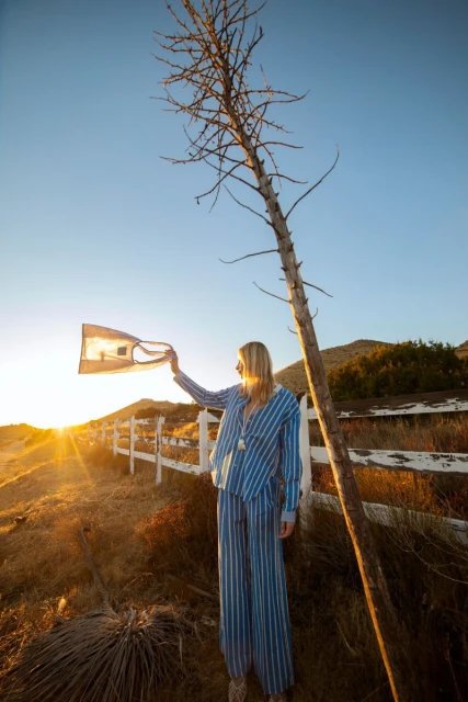 A woman in striped pajamas stands in a golden field at sunset, holding up fabric, beside a bare tree and a white wooden fence.