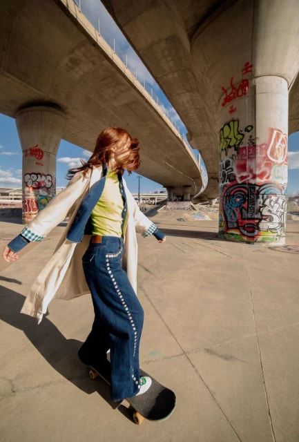 Woman rides a skateboard wearing denim jeans and jacket outdoors under a bridge with urban graffiti in bright daylight.