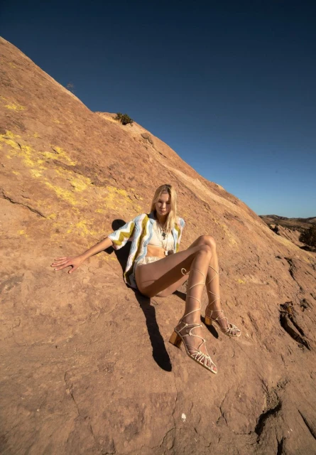 Fashion shot of a female model in a patterned shirt and gladiator sandals, sitting on a rocky landscape under a clear blue sky.