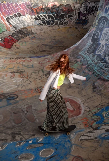 A woman in patterned pants and a white jacket rides a skateboard in a graffiti-covered skatepark, creating a dynamic lifestyle image.