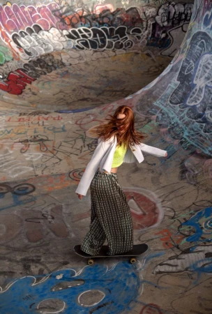 A woman in patterned pants and a white jacket rides a skateboard in a graffiti-covered skatepark, creating a dynamic lifestyle image.