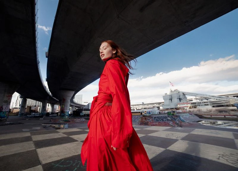 Woman in a red dress poses beneath a concrete overpass with graffiti and a checkered ground. Blue sky and clouds fill the background.