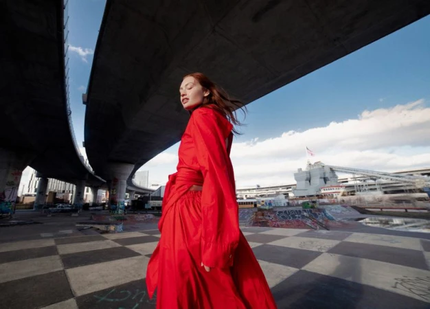 Woman in a red dress poses beneath a concrete overpass with graffiti and a checkered ground. Blue sky and clouds fill the background.
