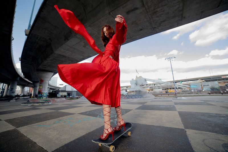 A woman in a red dress is skateboarding at an outdoor skatepark in front of a concrete bridge, wearing high heels and posing. 